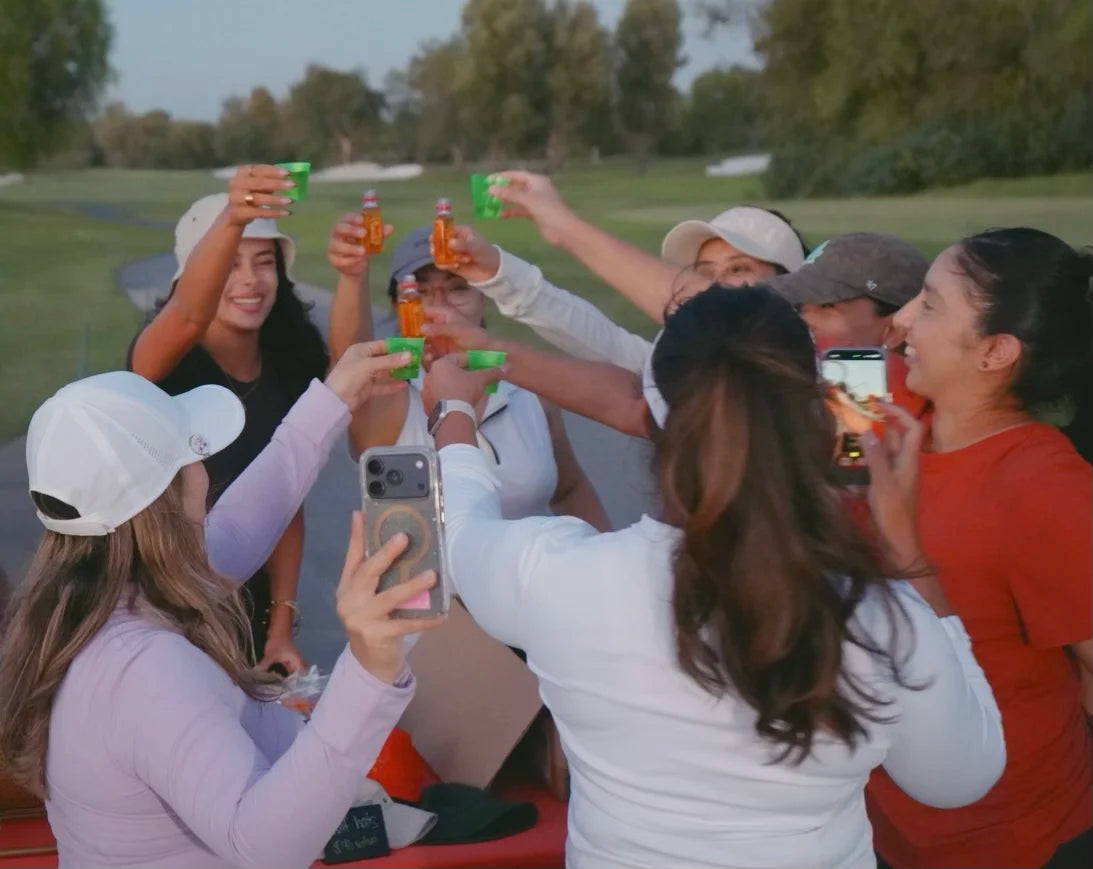 A group of friends celebrating on a golf course, raising green and amber bottles in a toast with their phones in the air. They are dressed in casual golf attire, including white and beige caps, with a green golf course in the background.