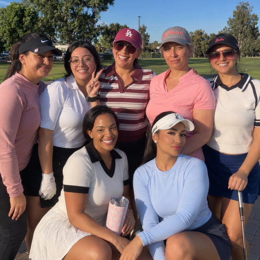 A group of seven women posing together outdoors on a golf course at sunset. They are wearing various casual and golf apparel outfits in pink, white, navy, and red, with trees and grass visible in the background.